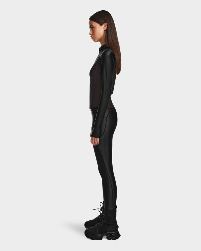 A woman stands in profile against a plain background, wearing Rudsaks CLARE - 8524519 Black fitted long-sleeve top and shiny stretch leggings, paired with chunky black boots. Her straight brown hair is worn down.