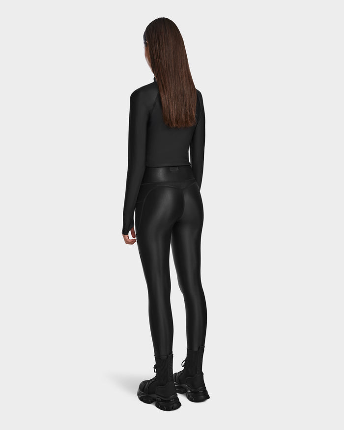 A woman with straight brown hair faces away, wearing the Rudsak CLARE - 8524519 Black fitted long-sleeve top and shiny stretch leggings that sculpt her silhouette, paired with chunky black shoes against a plain light gray background.