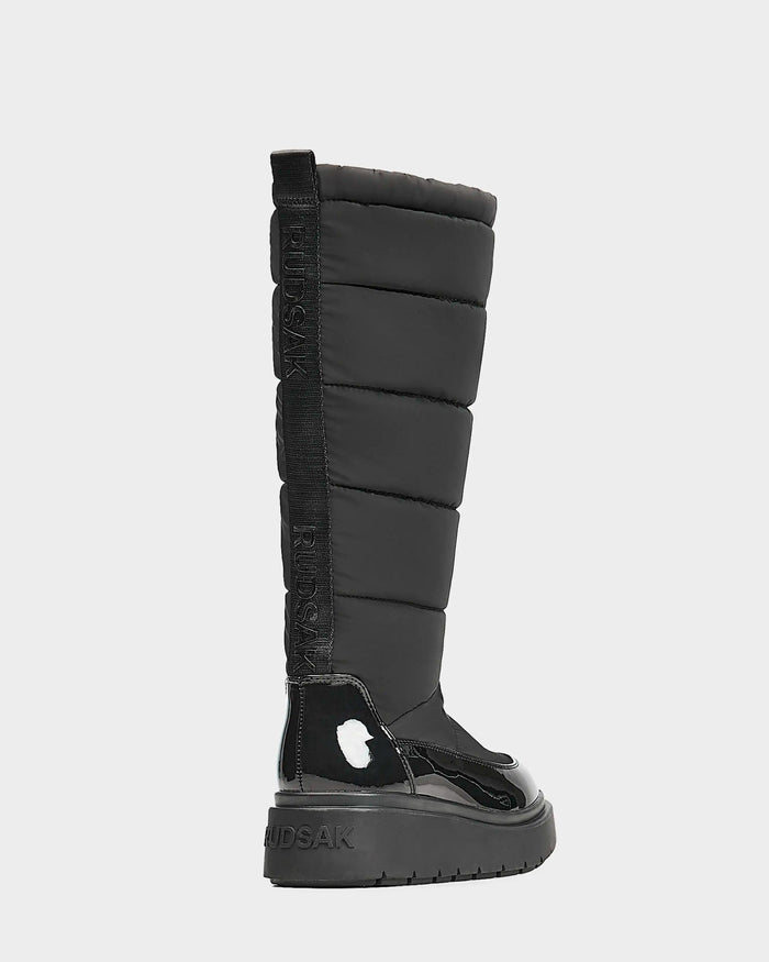 Side view of Rudsaks CLAUDE - L223506 Black womens tall quilted winter boot, featuring a padded shaft, glossy lower section, flat thick sole, and Rudsak branding on the back pull tab and heel. The boot is waterproof.