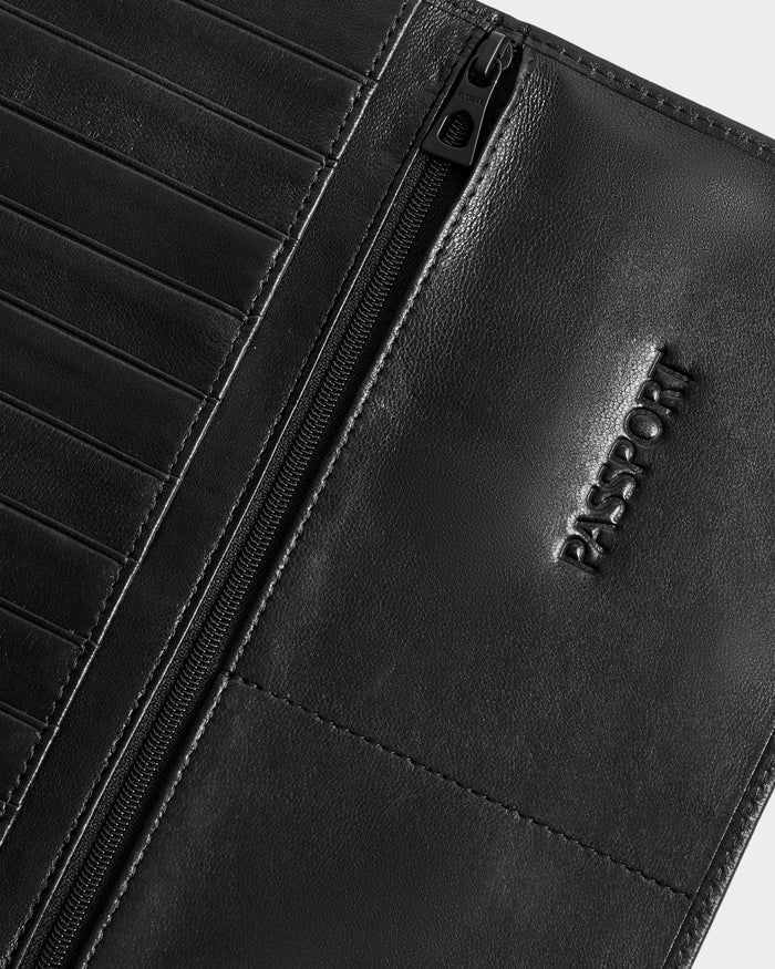 Close-up of the Rudsak CLEO - 9425522 Black unisex leather passport holder with an embossed “PASSPORT” label, zippered pocket, and card slots on a light gray background—an essential travel accessory.
