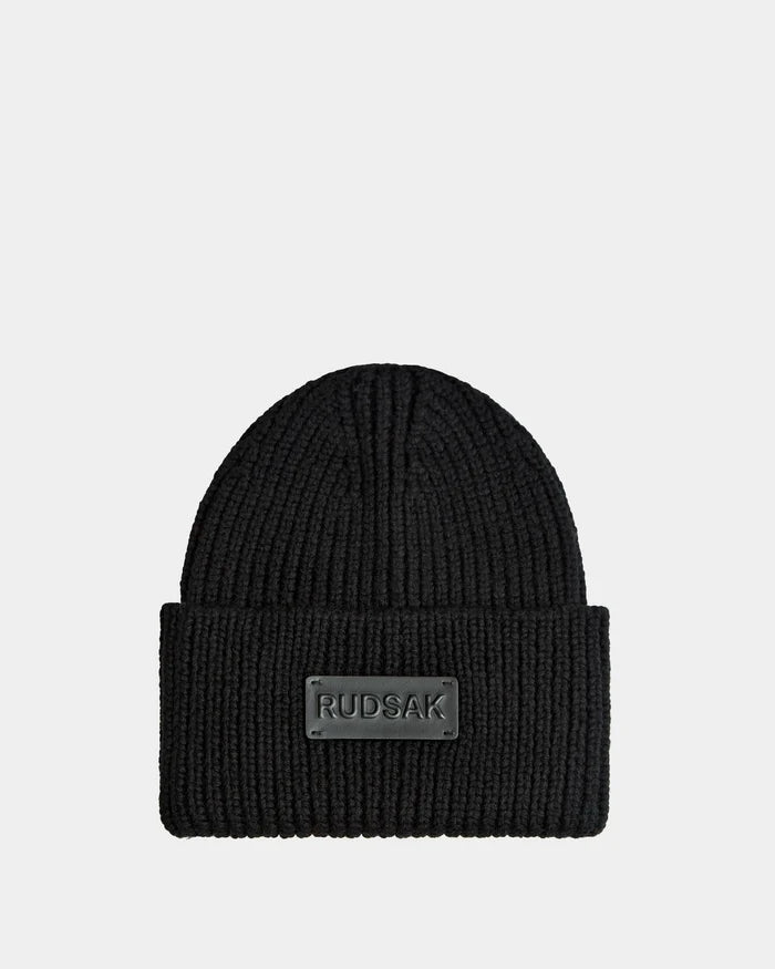 The COLIN - 9424518 Black by Rudsak is a unisex black ribbed knit beanie featuring a wide folded brim and a front rectangular patch with the brand name in raised letters.
