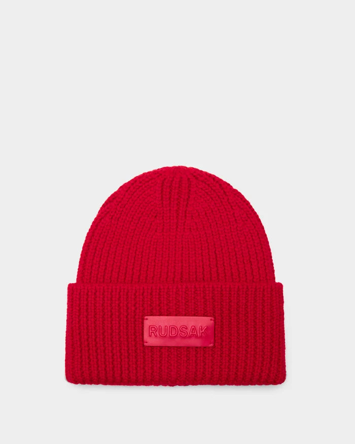 The Rudsak COLIN - 9424518 Flame Red is a bright red unisex ribbed beanie with a folded brim and a rectangular front patch featuring raised RUDSAK lettering. Displayed against a light gray background.