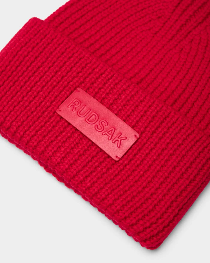 A close-up of the Rudsak COLIN - 9424518 Flame Red merino wool beanie with a ribbed texture, folded brim, and a rectangular rubber patch displaying the RUDSAK logo.