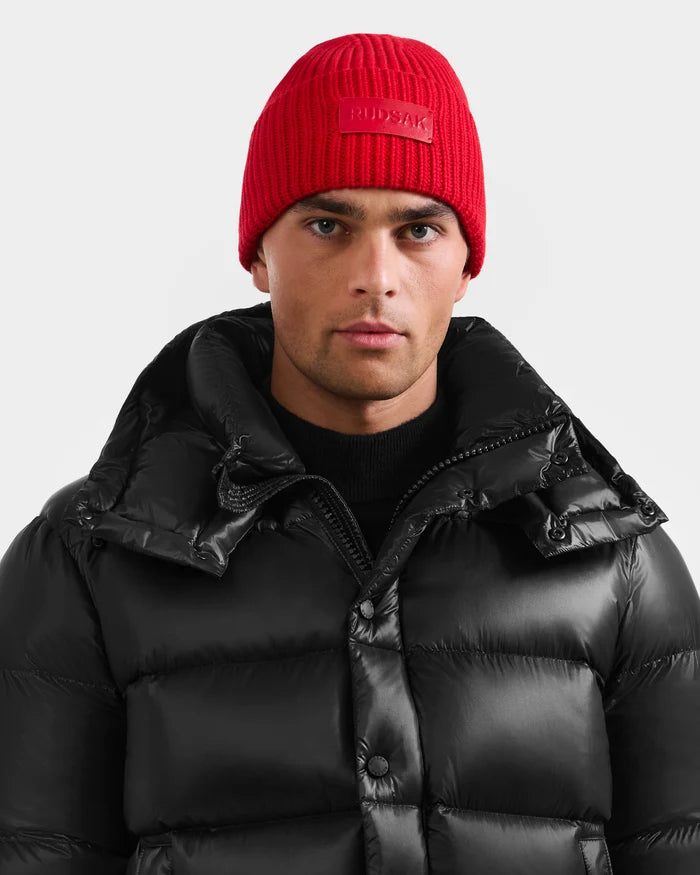 A man wears the Rudsak COLIN - 9424518 Flame Red beanie and a black puffer jacket with a high collar, standing against a plain light gray background.