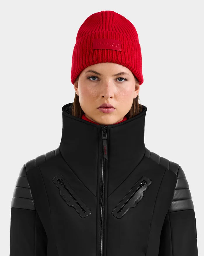 A young woman with straight brown hair faces forward against a light gray background, wearing the Rudsak COLIN - 9424518 Flame Red merino wool beanie and a high-collared black jacket with zipper details and leather accents.