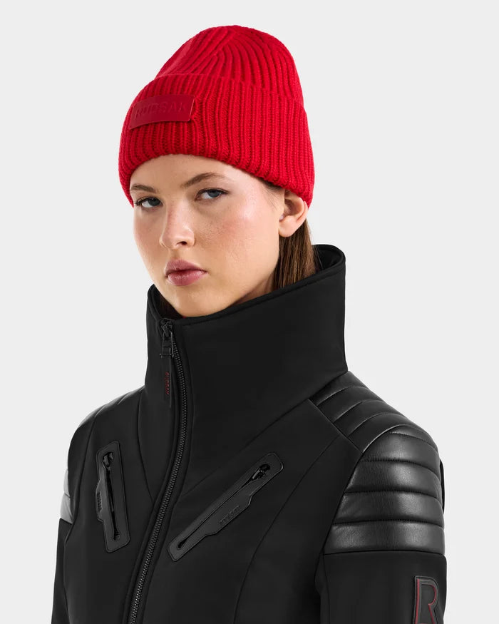 A woman wears the COLIN - 9424518 Flame Red merino wool beanie and a black Rudsak jacket with high collar and leather accents, looking confidently at the camera against a plain light background.