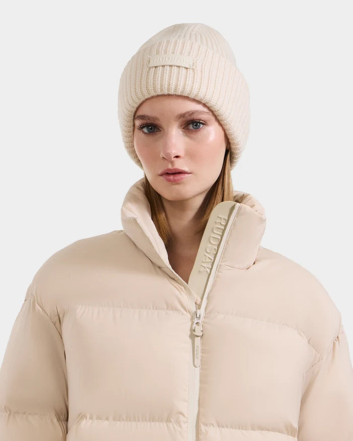 A woman with light brown hair and a neutral expression wears the Rudsak COLIN - 9424518 Vanilla cream beanie and matching puffer jacket with a high collar, both featuring subtle branding, against a plain background.