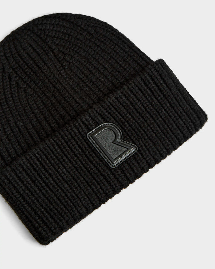 A close-up of the Rudsak CYBELLE - 9424504 Black ribbed knit beanie with a folded brim and raised black R patch, laid on a plain white background—a standout unisex winter accessory.