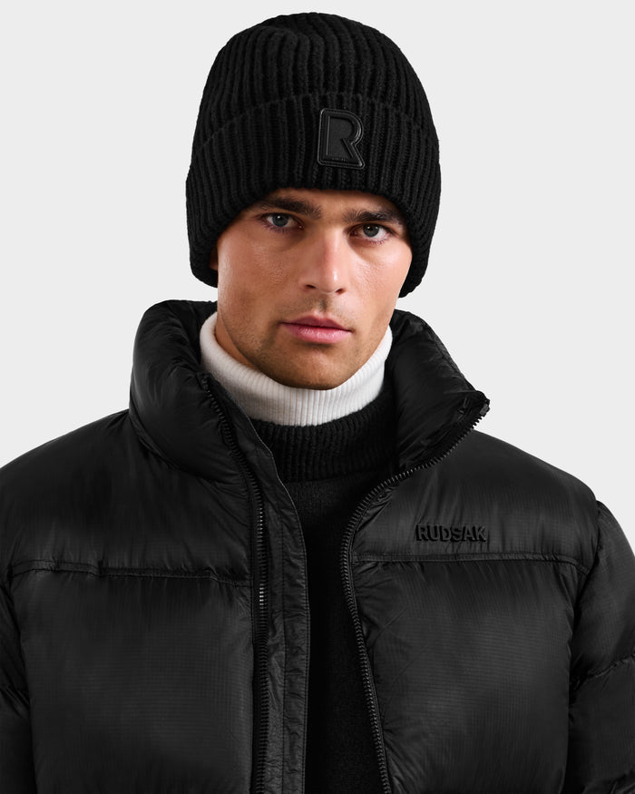 Wearing the unisex CYBELLE - 9424504 Black knit hat by Rudsak with a raised R logo, black puffer jacket, and black and white turtleneck, this person looks at the camera against a plain light background—an essential winter accessory.