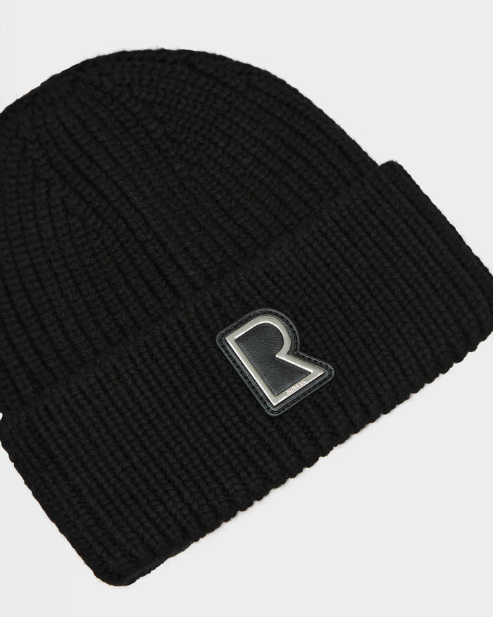 The CYBELLE - 9424504 Black/Silver by RUDSAK is a unisex black ribbed wool blend beanie with a folded brim and raised R logo patch on the front—an essential winter accessory.