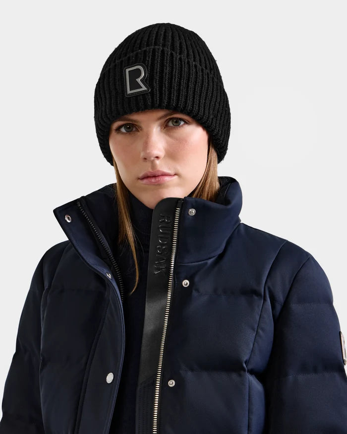 A person wearing Rudsak’s CYBELLE - 9424504 Black/Silver wool-blend beanie with an R logo, styled with a navy puffer jacket and black zip-up layer, stands against a plain light background.