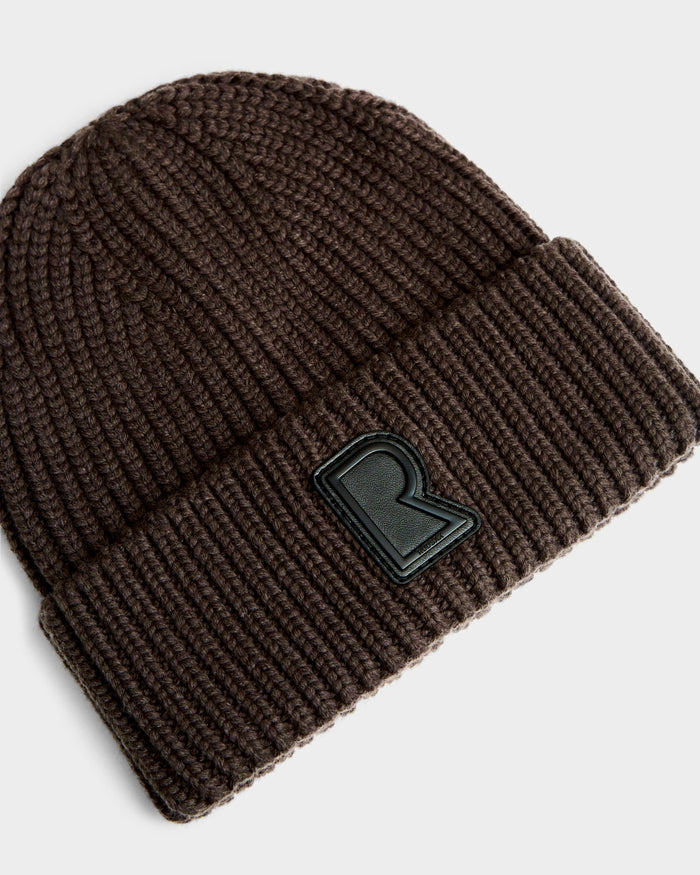 The Rudsak CYBELLE - 9424504 Espresso is a dark brown unisex winter beanie with a folded brim and a black raised R patch on the front, shown against a light gray background.