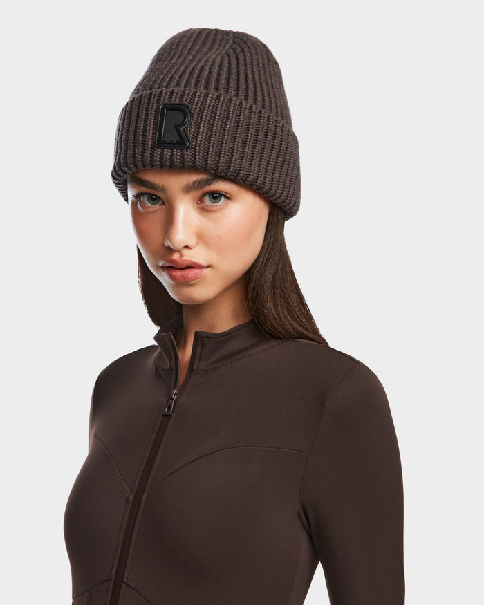 A woman models the CYBELLE - 9424504 Espresso beanie by Rudsak, featuring a dark brown wool blend and black R patch, paired with a matching zip-up top against a white background—a cozy winter accessory for effortless style.