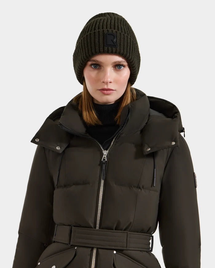 A woman wearing the Rudsak CYBELLE - 9424504 dark olive puffer coat with a belt and matching wool beanie stands against a plain background, looking directly at the camera to showcase this classic winter accessory.