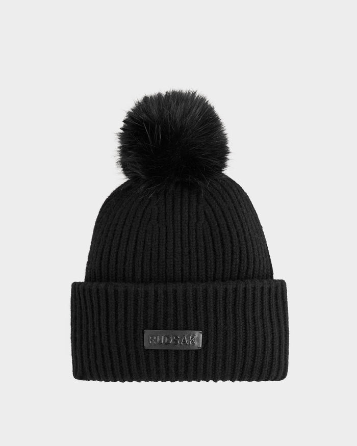 The DARA - 8425019 Black by Rudsak is a women's winter beanie with black ribbed knit, a folded brim, and a large faux fur pom pom. A rectangular patch with the Rudsak brand name adds a stylish touch to this cozy hat.