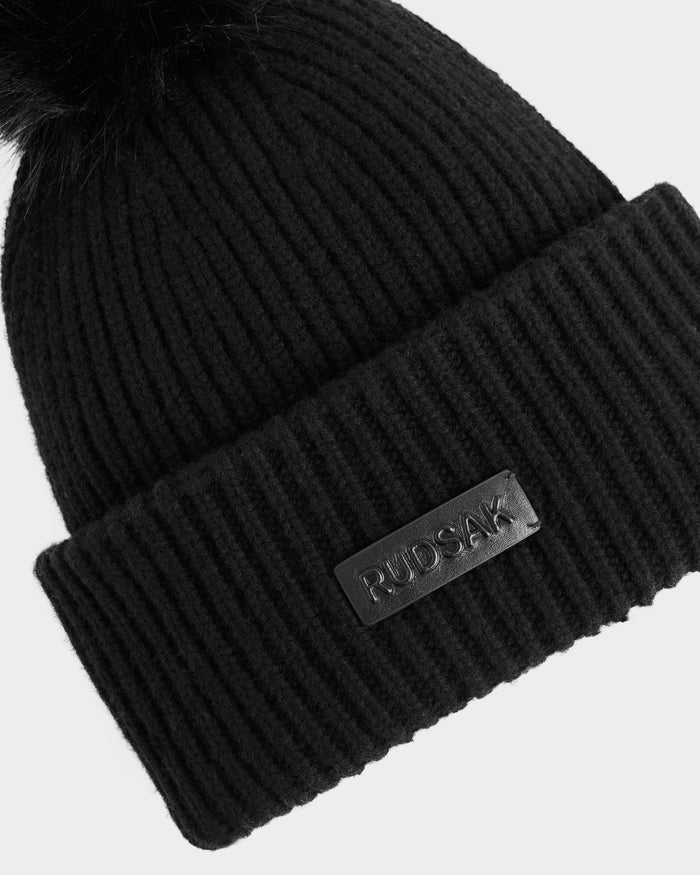 A close-up of the DARA - 8425019 Black beanie by Rudsak shows its ribbed knit, thick folded brim, a leather patch with embossed Rudsak logo, and a fluffy faux fur pom pom on top.