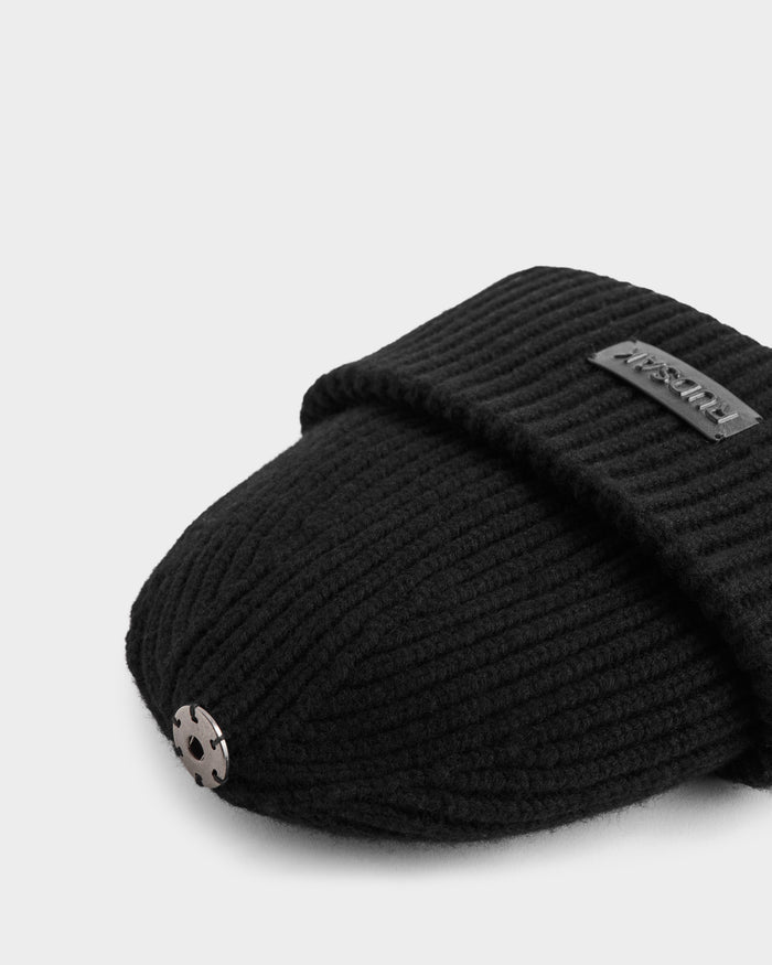 A close-up of the DARA - 8425019 Black slipper by Rudsak, featuring a ribbed black cashmere wool blend, a metallic circular stud on the toe, and a fabric label on the upper flap, set against a light background.