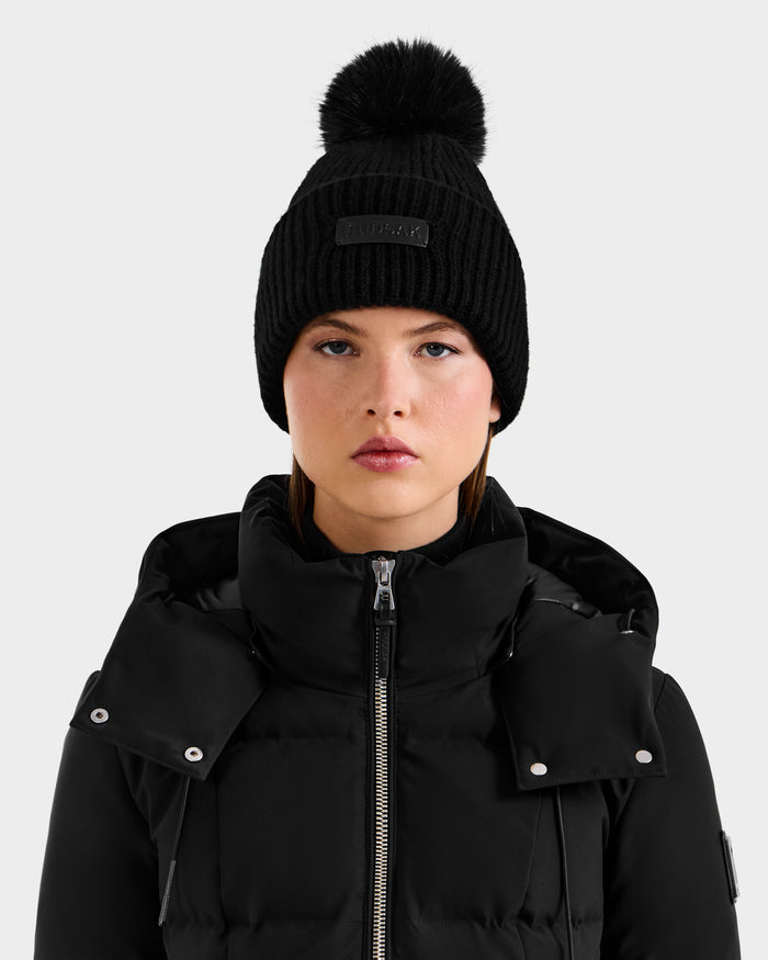 A woman wears the Rudsak DARA - 8425019 Black puffer jacket and a cashmere wool tuque with a faux fur pom pom, looking directly at the camera against a plain light gray background.
