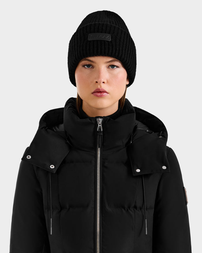 A woman wearing the Rudsak DARA - 8425019 black puffer jacket with a high collar and zipper, paired with a black winter hat, looks straight at the camera against a plain light gray background.