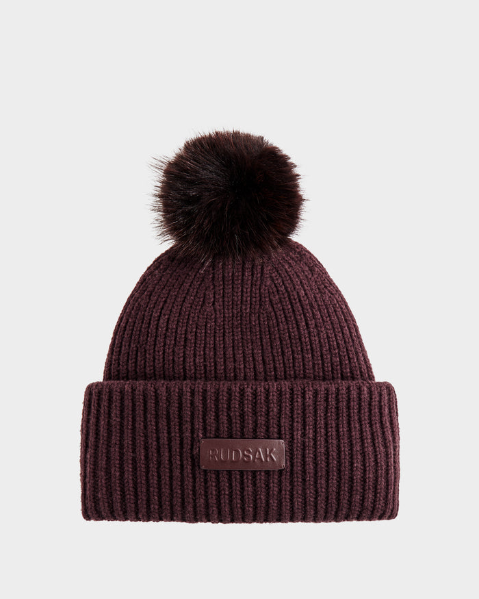 The Rudsak DARA - 8425019 Bordeaux is a women's dark burgundy ribbed knit beanie with a folded brim, faux fur pom-pom, and front brown leather patch embossed with RUDAK. Shown on a plain light gray background.