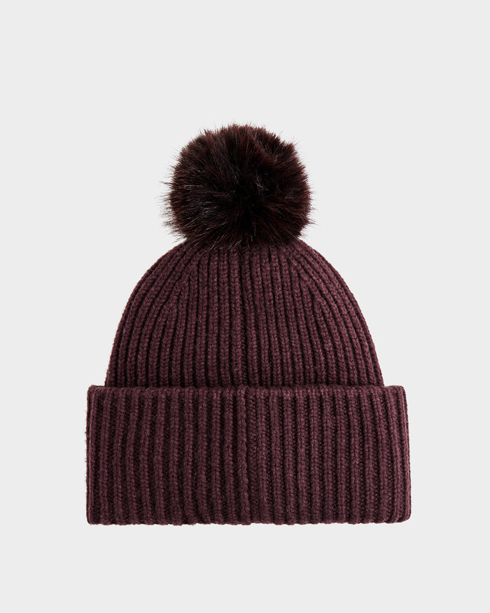 The DARA - 8425019 Bordeaux by Rudsak is a ribbed maroon knit cashmere wool tuque featuring a folded brim and a large faux fur pom pom, shown against a plain white background.