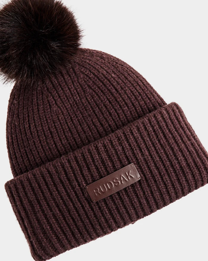 The DARA - 8425019 Bordeaux by Rudsak is a ribbed, dark brown women’s winter hat with a large faux fur pom pom, a folded cuff, and a leather Rudsak patch.