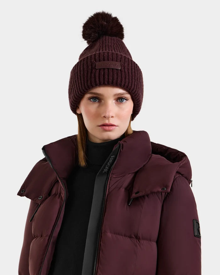 A woman in the DARA - 8425019 Bordeaux tuque by Rudsak, featuring a faux fur pom pom, pairs it with a matching maroon puffer jacket over a black turtleneck, standing against a plain light background.