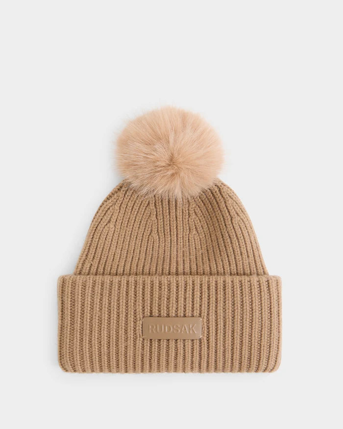 The DARA - 8425019 Camel by Rudsak is a women’s tan ribbed knit tuque made from a cozy cashmere wool blend. It features a large, fluffy faux fur pom pom on top and a folded brim with a rectangular patch embossed with the Rudsak logo, shown against a plain white background.