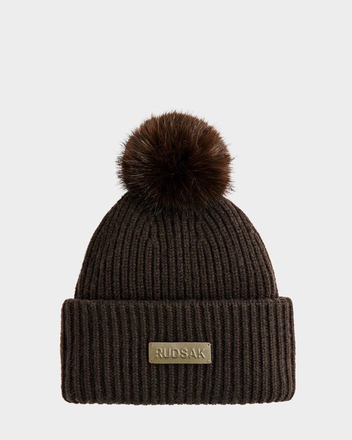 The DARA - 8425019 Dark Olive beanie by Rudsak features ribbed knit fabric, a large faux fur pom pom, and a folded brim with a gold Rudsak label, all shown against a plain white background.