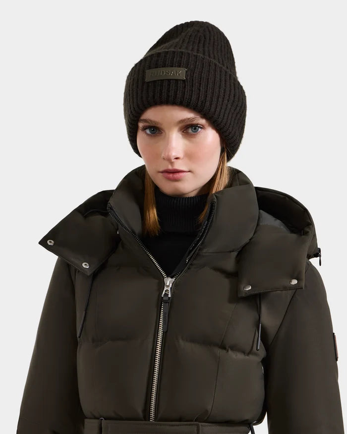 A woman wears the Rudsak DARA - 8425019 dark olive puffer jacket and a ribbed knit women's winter hat with a logo, looking straight ahead against a plain light background.