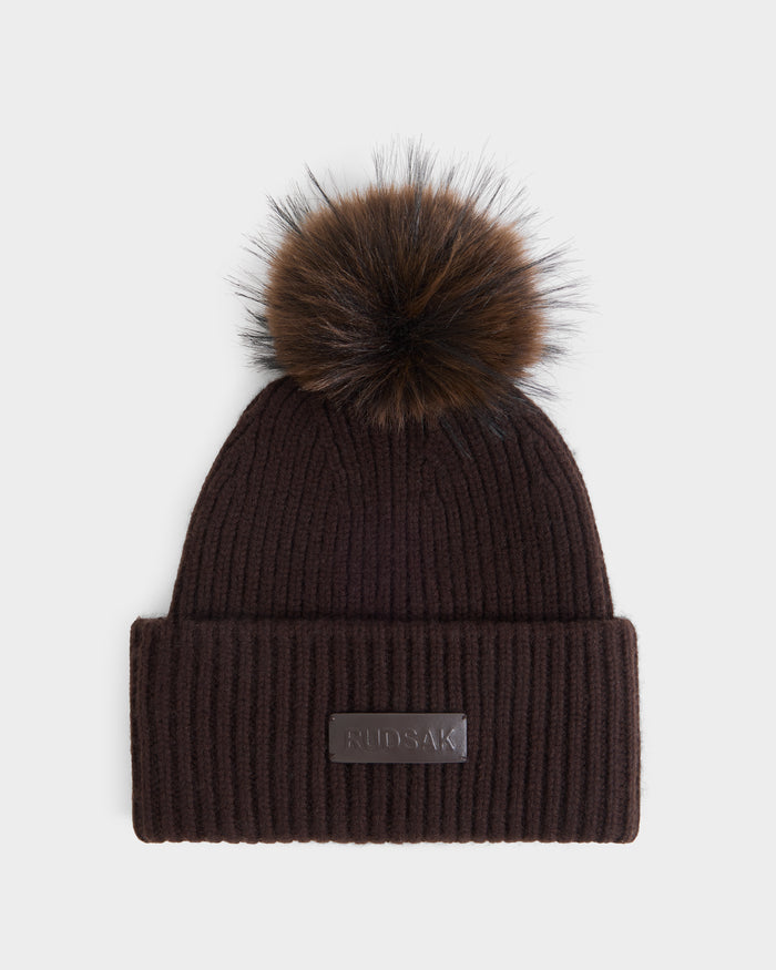 The DARA - 8425019 Espresso by Rudsak is a dark brown ribbed knit women’s winter hat, featuring a wide folded brim, a large faux fur pom-pom on top, and a rectangular leather patch with embossed branding on the front.