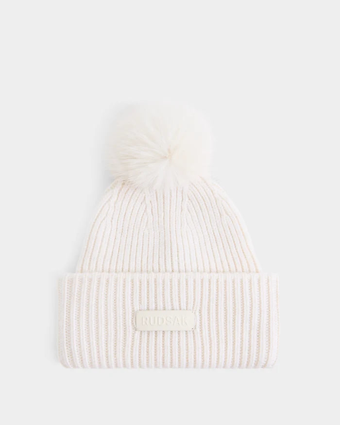The DARA - 8425019 Vanilla by Rudsak is an off-white ribbed knit women’s tuque featuring a folded brim, a fluffy faux fur pom pom on top, and a signature RUDSAK label on the front. Crafted from a luxurious cashmere wool blend, this hat is shown against a plain white background.