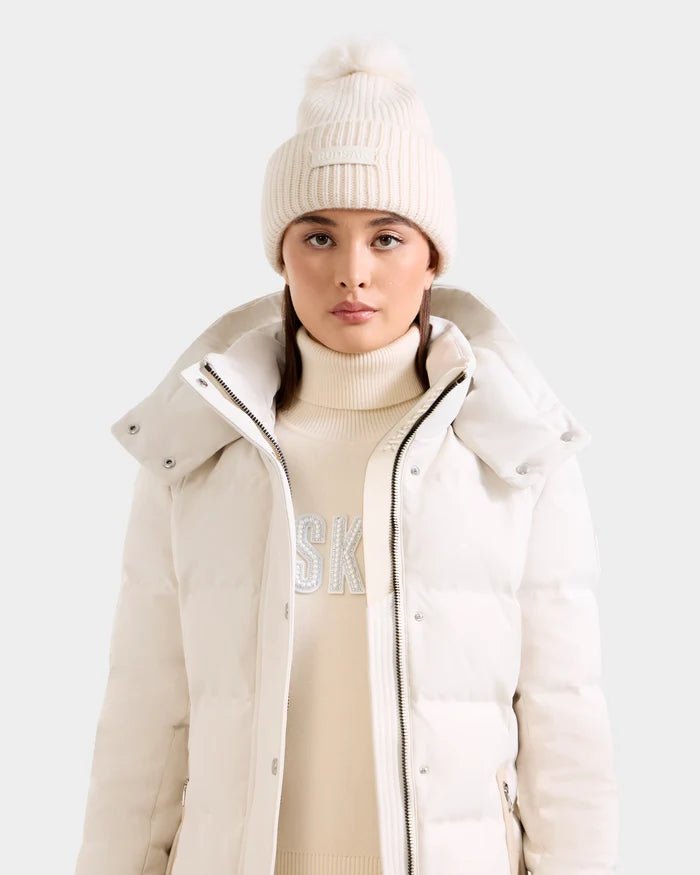 A woman wears the Rudsak DARA - 8425019 Vanilla cream cashmere wool tuque with faux fur pom pom, a light puffer jacket, and a cream turtleneck sweater against a plain light background, looking at the camera.