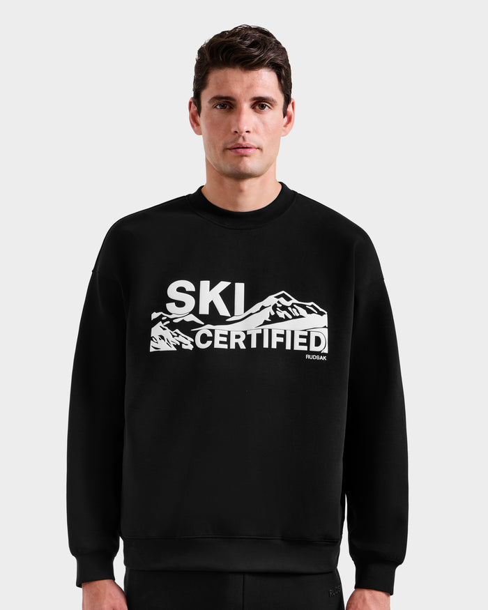 A man wears the Rudsak DECLAN - 6525648 black men’s crewneck sweatshirt with a white mountain graphic and "SKI CERTIFIED" text, standing against a plain light background to highlight this stylish ski piece.