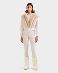 A woman faces forward wearing the Rudsak DELLA - 8525668 Vanilla off-white faux fur hoodie with a large beige collar, matching fitted pants, and cream snow boots for an effortlessly chic après-ski look against a plain light background.
