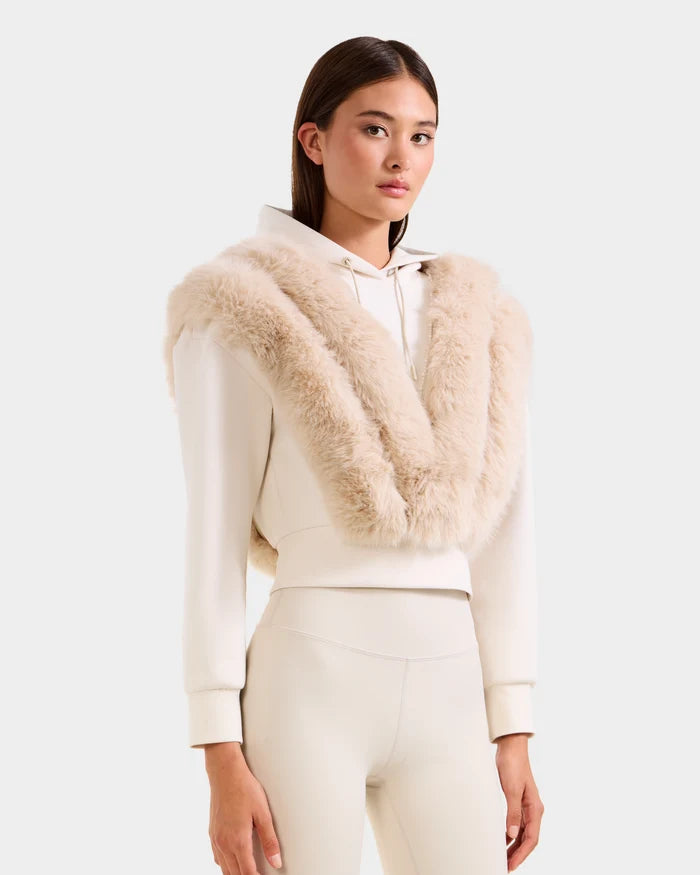 A woman wearing the Rudsak DELLA - 8525668 Vanilla cream après-ski pullover with a faux fur shawl draped over her shoulders stands against a plain white background.