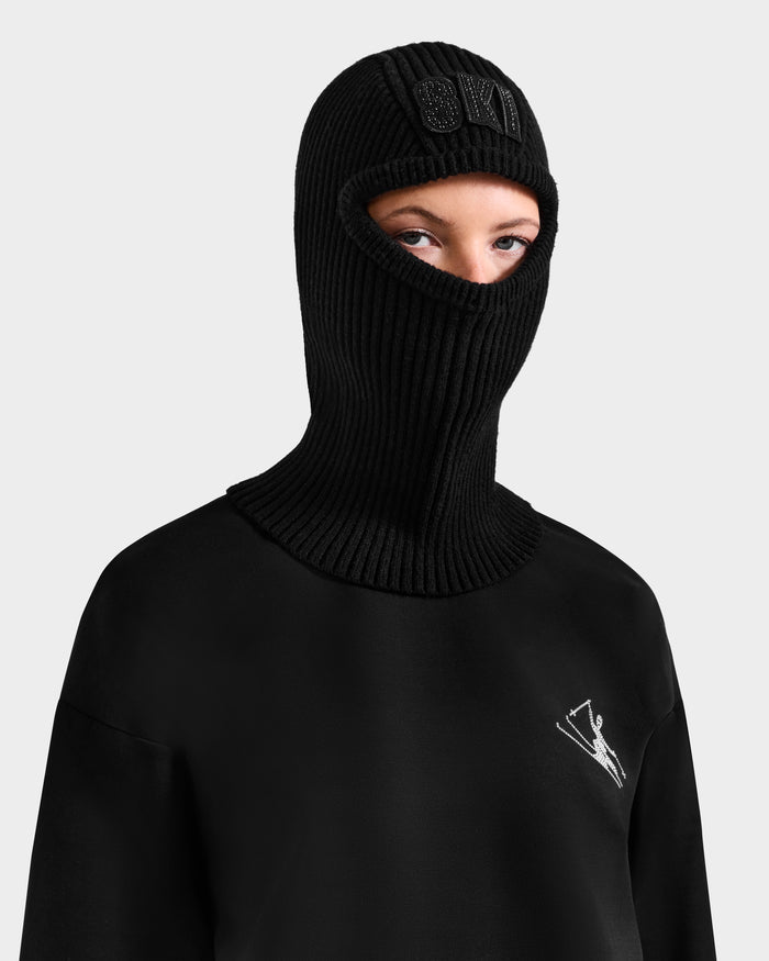 A person wears the Rudsak DIXIE - 8425551 Black women's knit balaclava and a black sweatshirt with a small white chest graphic, standing against a light gray background—an ideal look to update your winter wardrobe.