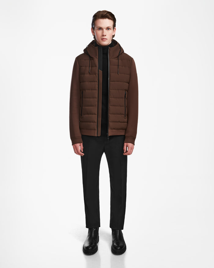 A person faces forward against a plain white background, wearing Rudsaks ELIAS - M124002 Espresso wind- and water-resistant jacket over a black top, with black trousers and boots.