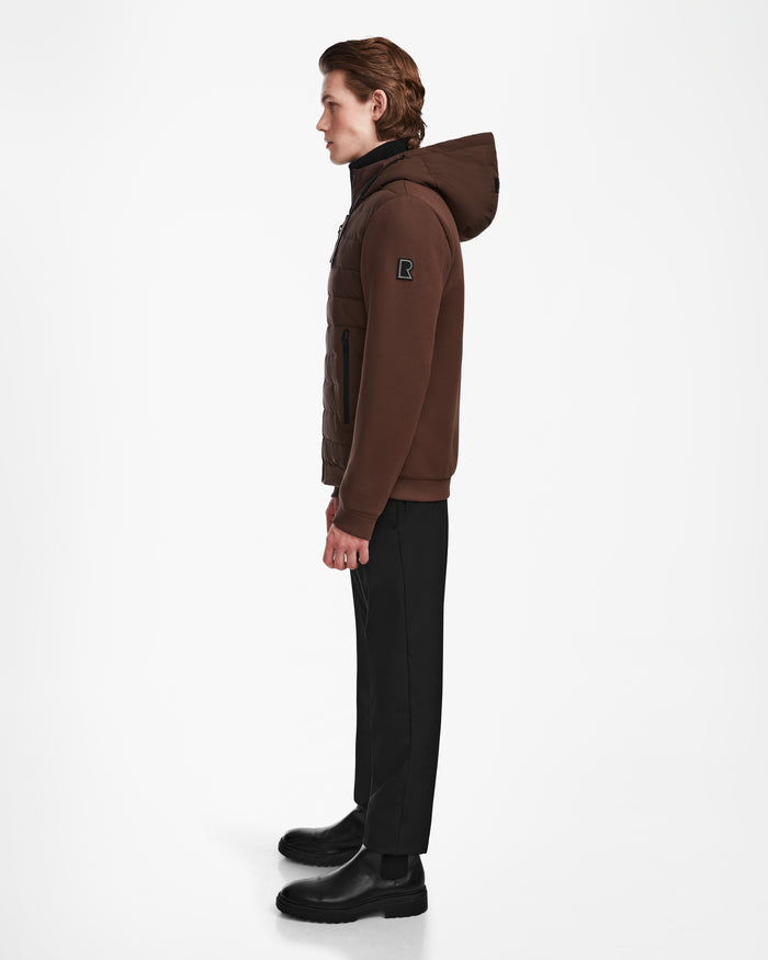 A person with brown hair stands in profile against a white background, wearing the Rudsak ELIAS - M124002 Espresso men’s quilted jacket with a hood, paired with black pants and boots.