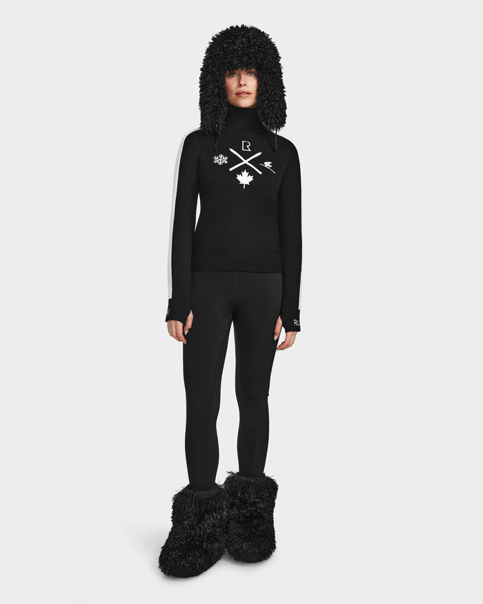 A person stands facing forward in the Rudsak ELIZABETH - 8524623 Black womens ski turtleneck with white symbols, paired with black leggings and matching furry accessories, all set against a plain white background. Ideal for any winter wardrobe.