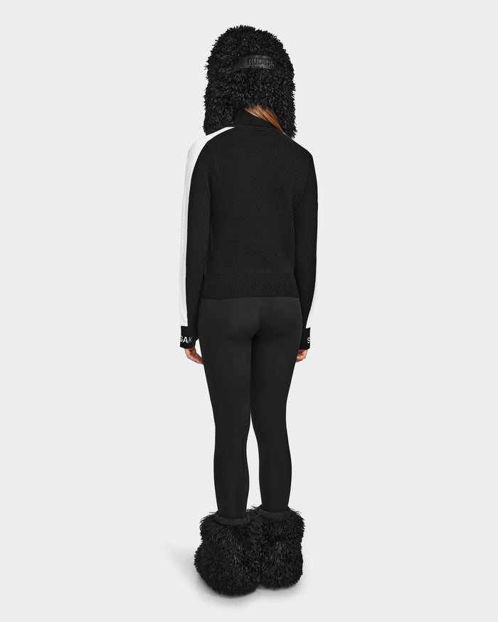 Against a plain white background, a person faces away wearing Rudsaks ELIZABETH - 8524623 Black hat with its fluffy textured material, paired with a black and white ski turtleneck, black leggings, and matching black fluffy boots for chic après-ski style.