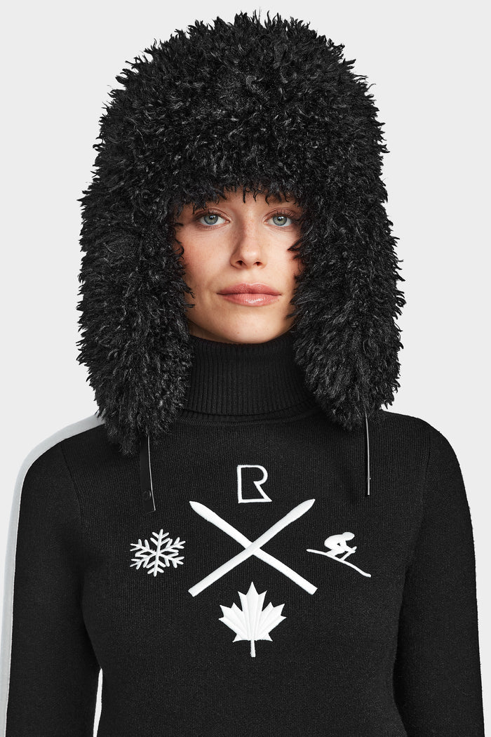 Dressed for après-ski, a woman wears the Rudsak ELIZABETH - 8524623 Black: a cozy, fuzzy black hat and a women’s ski turtleneck featuring white graphics—snowflake, ski poles, skier, maple leaf, and the letter R.