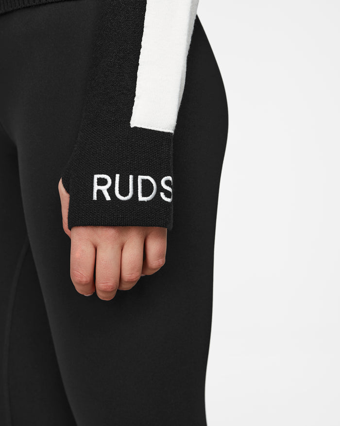 Wearing the Rudsak ELIZABETH - 8524623 Black leggings and a womens ski turtleneck with a white vertical stripe and RUDS text on the sleeve, this après-ski look is effortlessly chic, hand relaxed at their side.