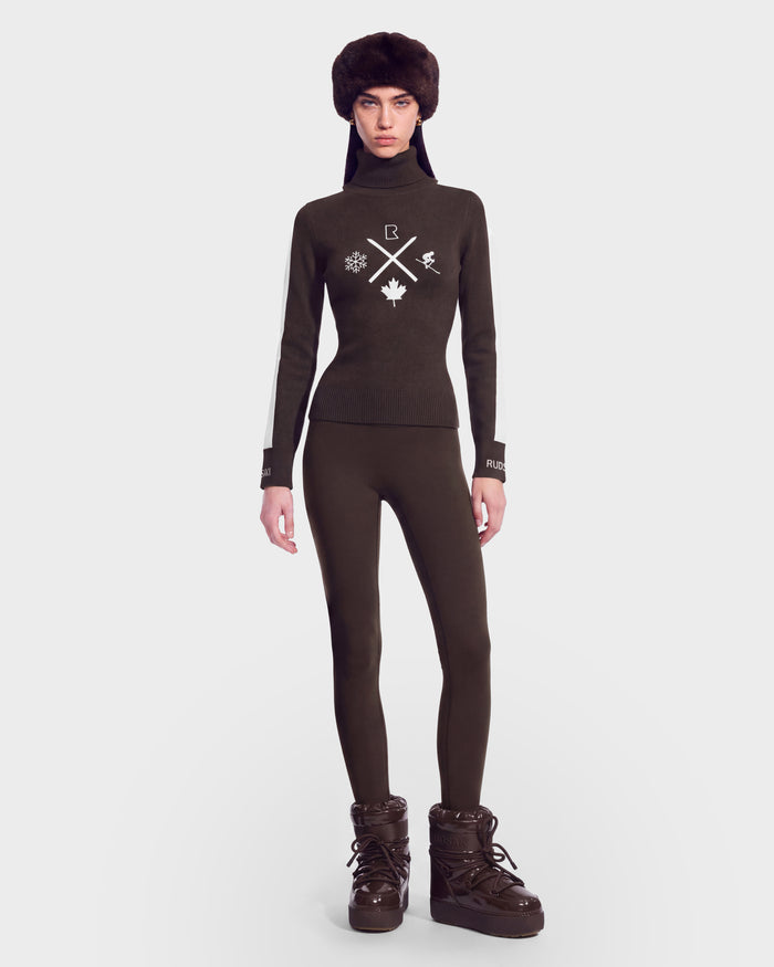 Wearing the ELIZABETH - 8524623 Espresso sweater by Rudsak, this person pairs the dark, fitted knit—adorned with classic winter motifs and turtleneck styling reminiscent of women’s ski apparel—with matching leggings, furry boots, and a fur hat. Set against a plain white background, the look radiates effortless chic for any winter wardrobe or après-ski gathering.