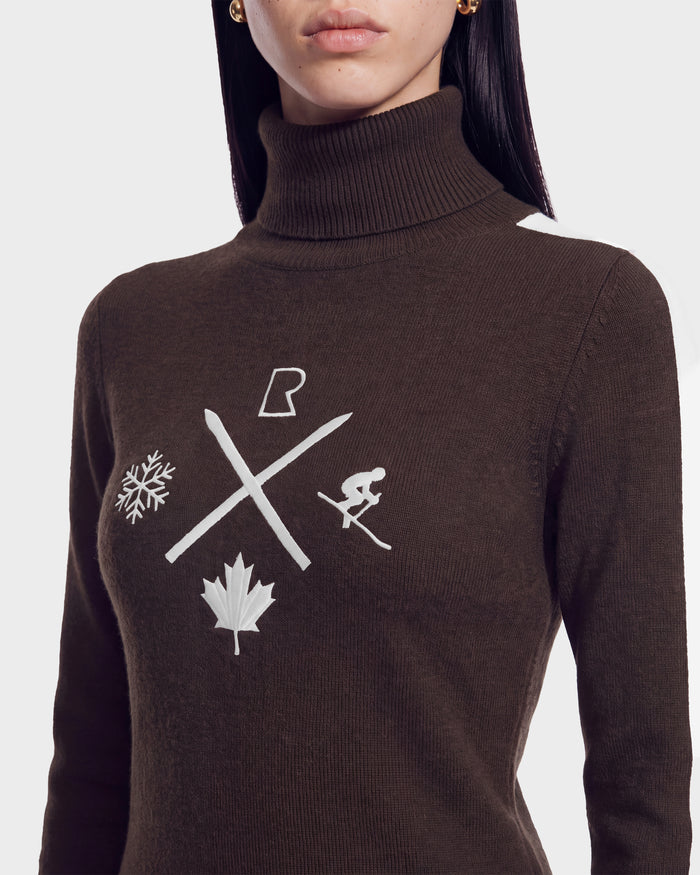 A woman in the ELIZABETH - 8524623 Espresso women’s ski turtleneck sweater by Rudsak, featuring a dark design with white symbols—a snowflake, maple leaf, skier, X, and a stylized letter R—making it an essential piece for any winter wardrobe or après-ski ensemble.