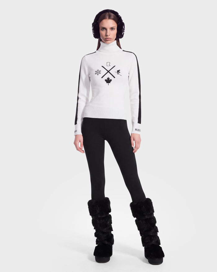 A woman stands facing forward against a plain white background, wearing plush earmuffs, black leggings, tall black fur boots, and the Rudsak ELIZABETH - 8524623 Optic White women’s ski turtleneck sweater featuring black snowflake designs—a perfectly cozy choice for any winter wardrobe or après-ski moment.