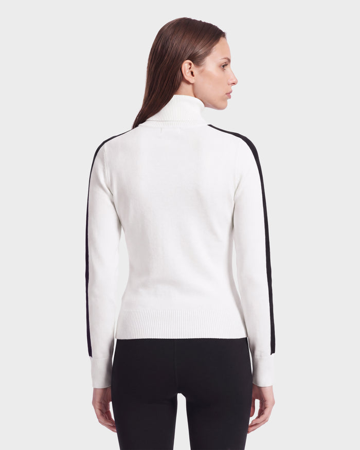 A woman with straight brown hair is seen from behind against a plain light background, wearing the ELIZABETH - 8524623 Optic White women’s ski turtleneck sweater by Rudsak, which features black stripes on the sleeves and is paired with black pants—an effortlessly stylish addition to any winter wardrobe.