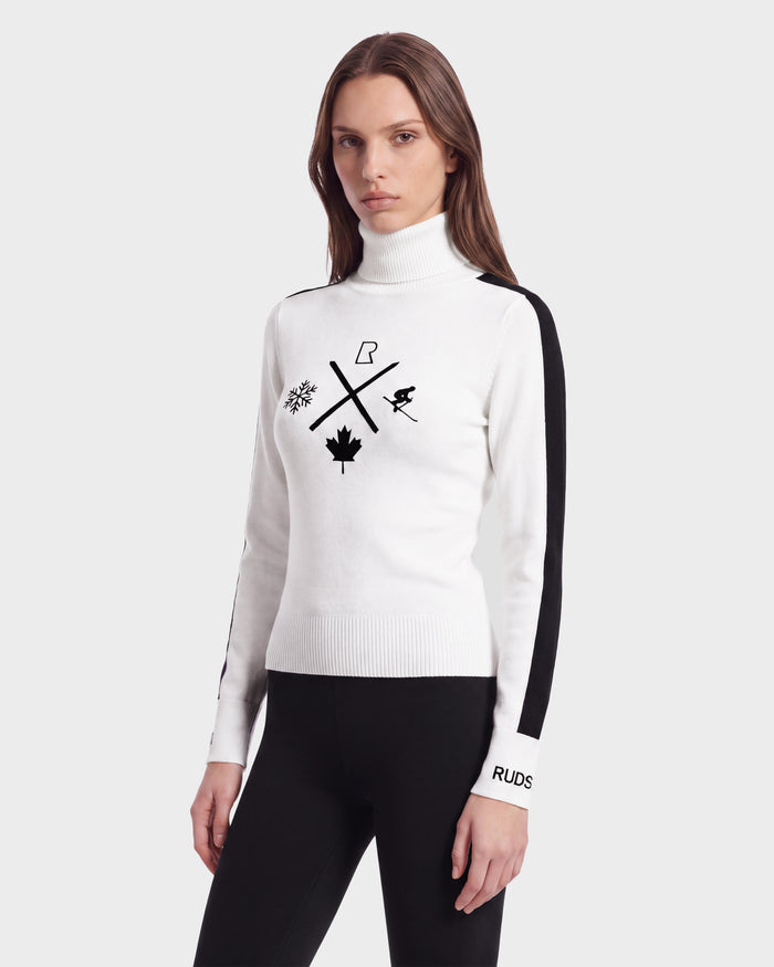 A woman models the Rudsak ELIZABETH - 8524623 Optic White women’s ski turtleneck, featuring chic black symbols and bold black side-striped sleeves, paired with sleek black pants—making it an ideal addition to your winter wardrobe—against a plain light background.