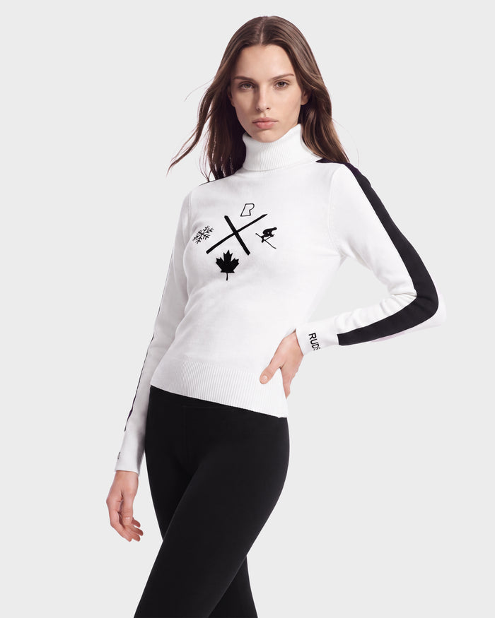 A woman stands with one hand on her hip against a plain light background, wearing the Rudsak ELIZABETH - 8524623 Optic White après-ski inspired women’s ski turtleneck sweater with black symbols and black side-striped pants, embodying the perfect addition to any winter wardrobe.