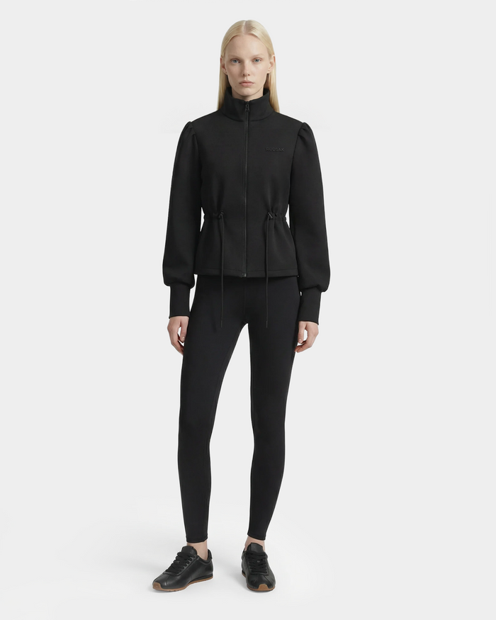 A woman with straight blonde hair stands against a white background, wearing the Rudsak ELODIE - 8526039 Black zip-up sweater jacket. She faces forward in black leggings and sneakers, highlighting this lightweight stretch cotton-blend layer.